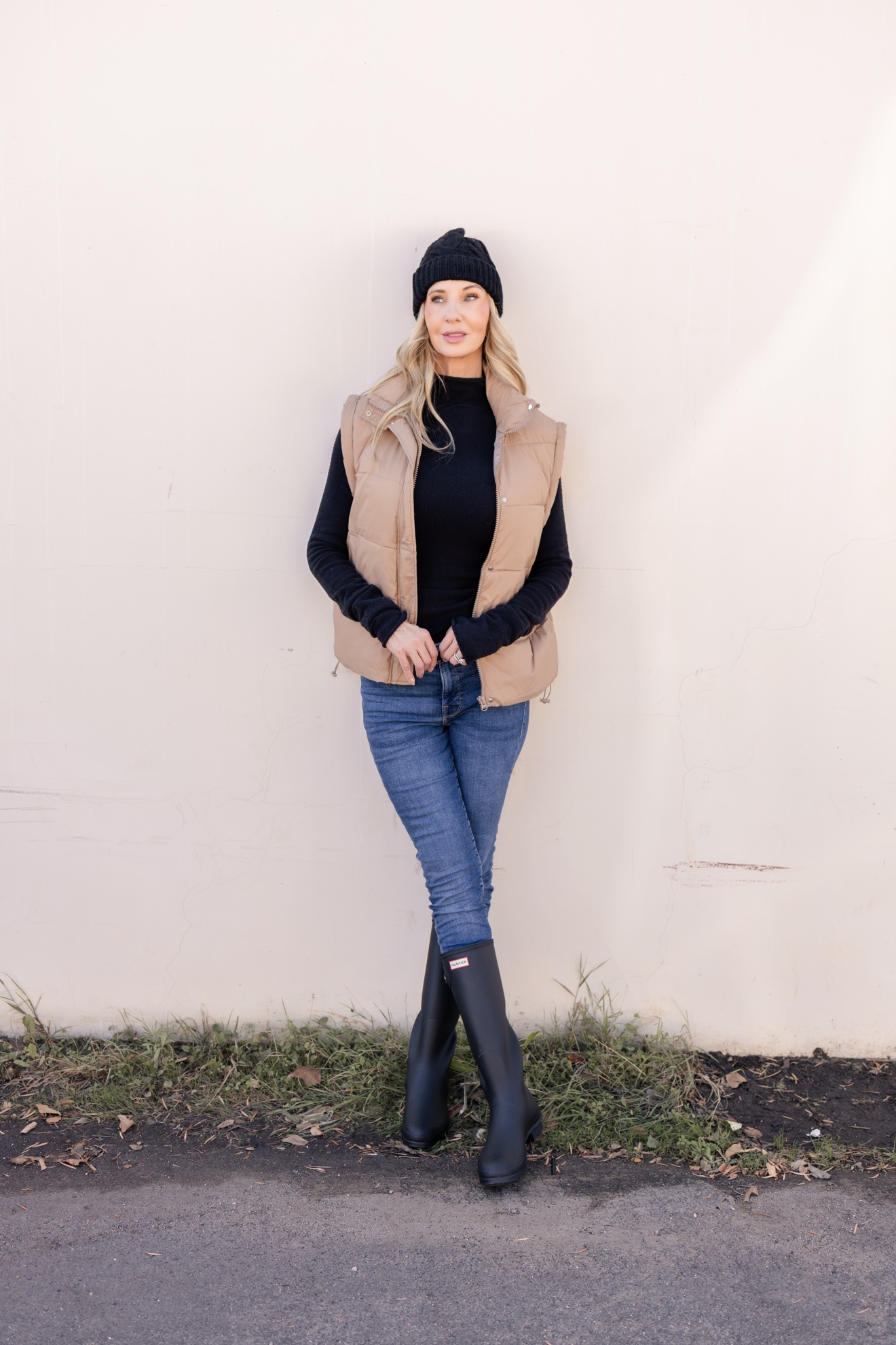 Ris straight-on in a tan puffer vest, skinny jeans, and tall boots against a cream wall