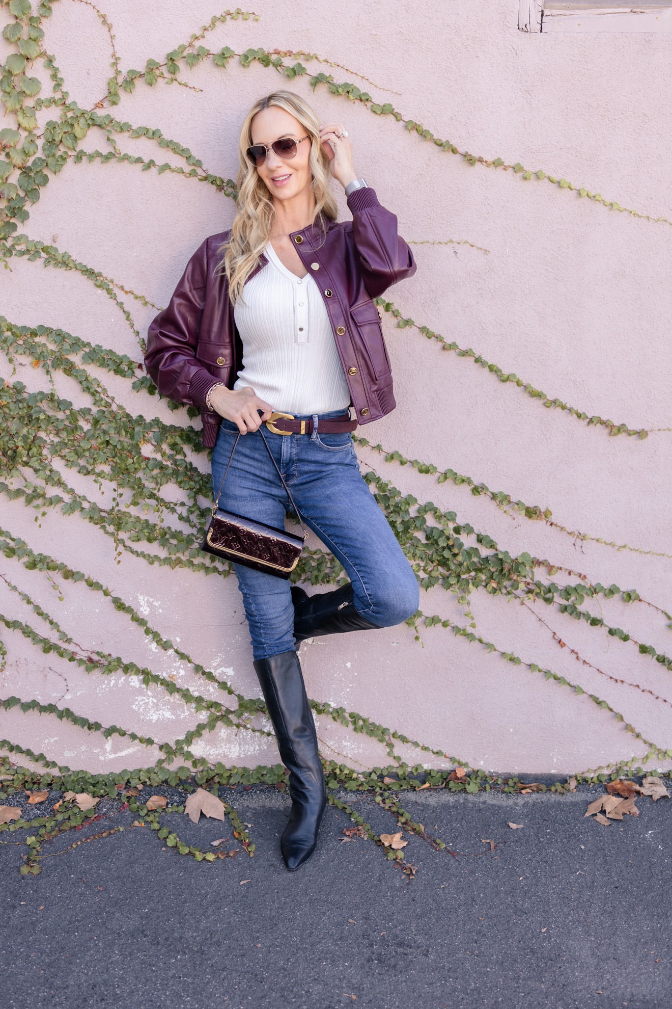 Ris in a burgundy leather jacket, jeans, and tall boots at an ivy wall