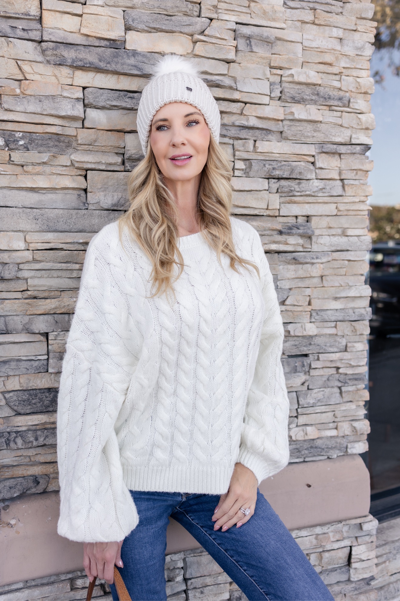 Ris in a white cable-knit sweater and cream beanie against a stone wall